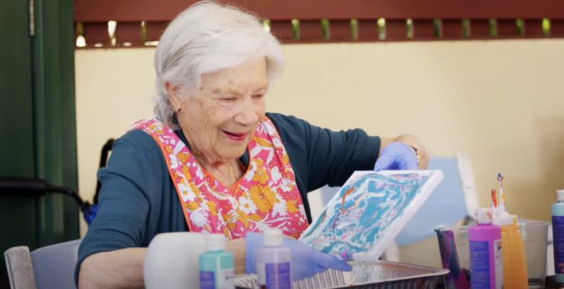 Anglicare client, Valerie, painting a canvas outside while smiling