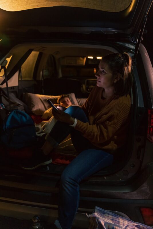 Woman experiencing homelessness sitting in boot of her car at night with young child