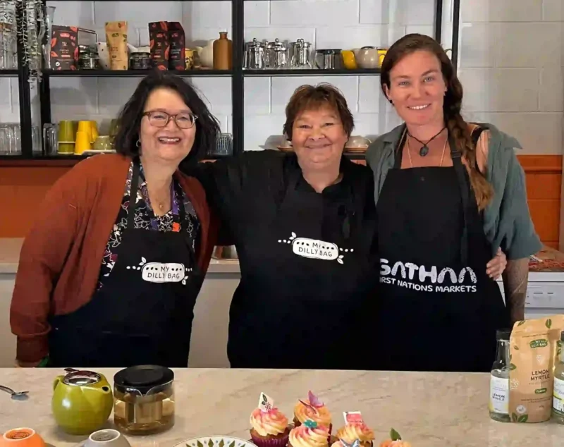 Three ladies standing behind a kitchen bench, smiling at the camera. There are cups and saucers on the bench and cupcakes too.