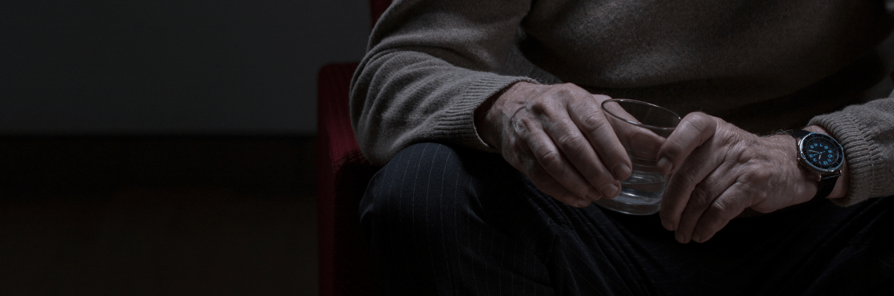 A person wearing a beige jumper and dark trousers sits in a dimly lit room, holding an empty glass with both hands—a quiet moment reflecting alcohol awareness. Only the hands and torso are visible; the person wears a wristwatch.