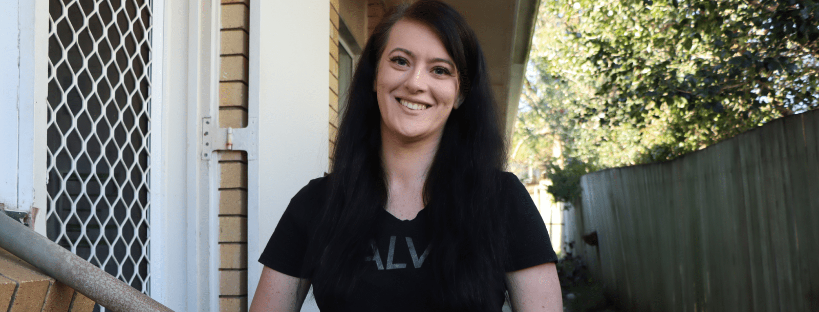 A woman with long dark hair stands outside near a house, smiling at the camera. Wearing a black T-shirt and holding a handrail, she looks content—far from sleeping rough—with trees and a green fence in the background.