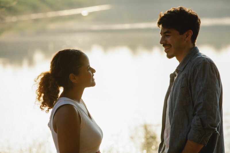 A woman and a man stand facing each other outdoors near a body of water, smiling and talking in warm, natural sunlight, their assertive communication evident as they engage openly. The background is softly blurred with reflections on the water.