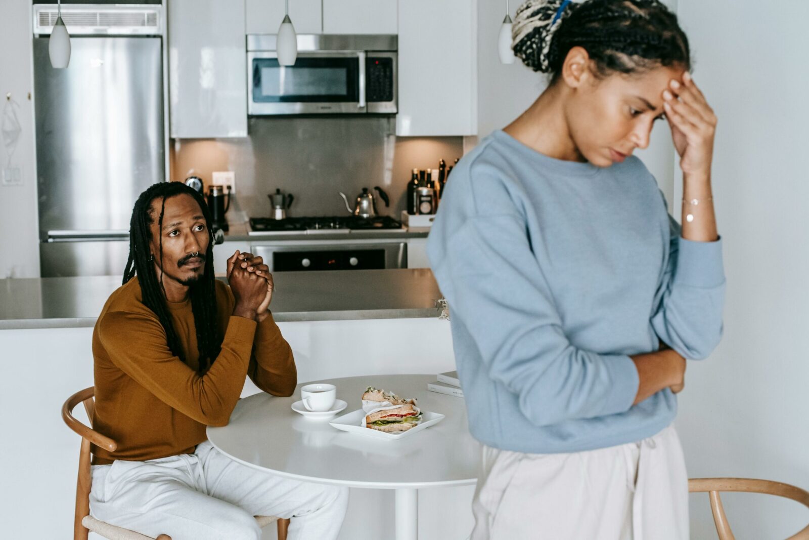 A man sits at a kitchen table with a sandwich and coffee, looking concerned, while a woman stands in the foreground with her head in her hand, appearing upset. The scene suggests a relationship breakdown between them.