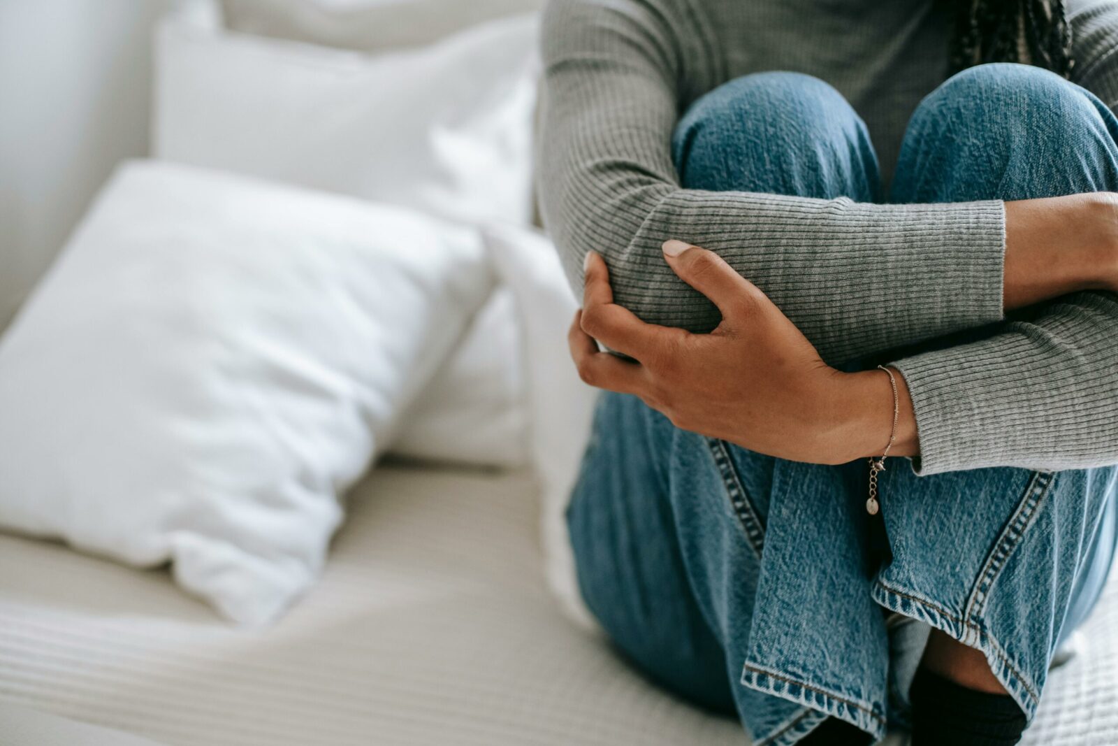 A person wearing a grey jumper and blue jeans sits on a bed, hugging their knees to their chest—an image that quietly reflects the weight of suicidal thoughts. White pillows and bedding are visible in the background; the person's face is not shown.
