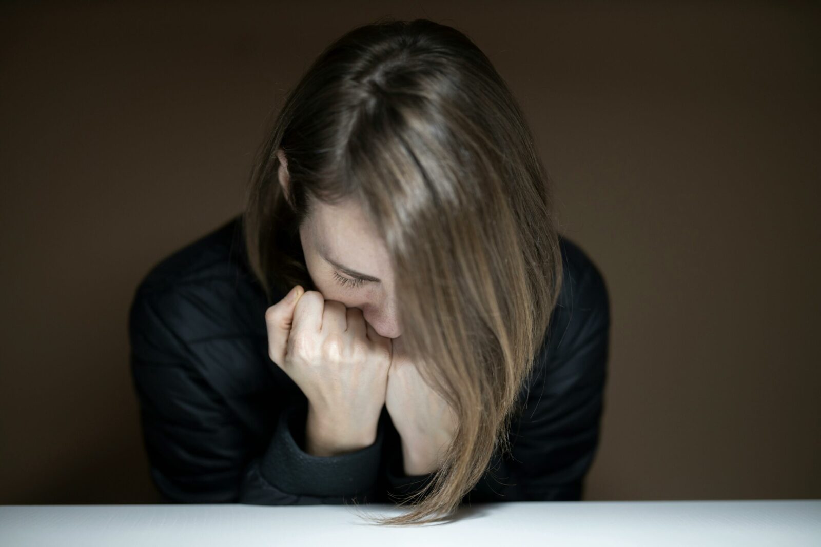 A person with long hair, dressed in a dark jacket, sits with their head bowed and fists pressed against their face, appearing thoughtful or showing signs of depression against a plain background.