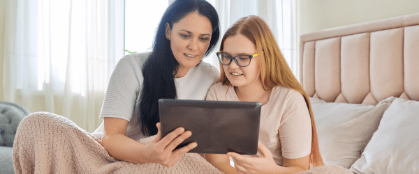 A woman and a girl with glasses sit on a bed under a duvet, smiling at a tablet together in a bright, cosy bedroom—sharing moments that show what makes a good foster carer.