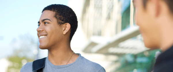 A young man with short curly hair smiles whilst standing outdoors, wearing a grey shirt and a rucksack—possibly meeting foster parents of adolescents. Another person is blurred in the foreground, with a modern glass building in the background.