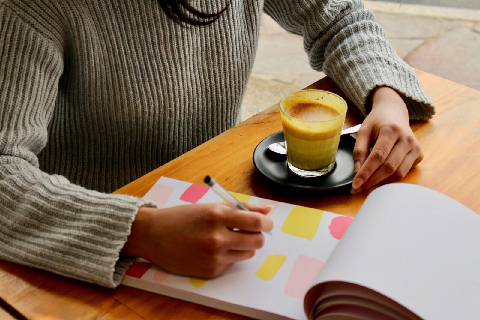 Person in a grey jumper writing about goal setting in a colourful notebook at a wooden table, with a glass of yellow latte on a black saucer beside them.