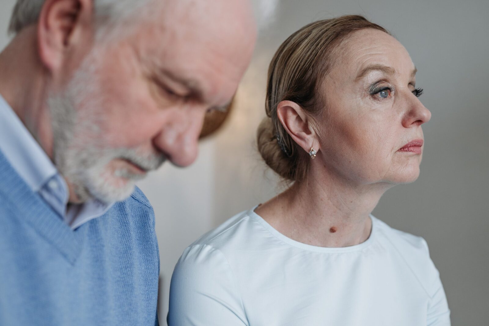 An older man and woman sit side by side indoors. The man looks downward with a serious expression, whilst the woman gazes off to the side, both appearing deep in thought as they quietly process grief and loss.