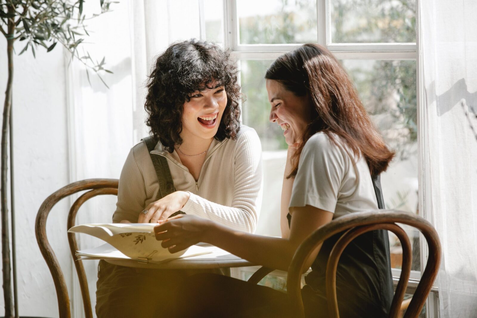 Two young women sit together at a small table by a sunny window, sharing a moment of intimacy as they smile and laugh over an open book or magazine. The scene appears light-hearted and friendly.