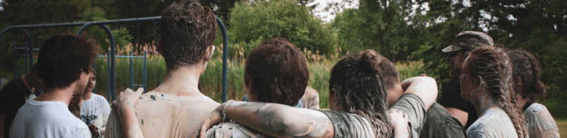 A group of people, celebrating youth day, stand close together outdoors with arms around each other's shoulders, covered in mud—suggesting a playful or team-building activity. Trees and grassy plants fill the background.