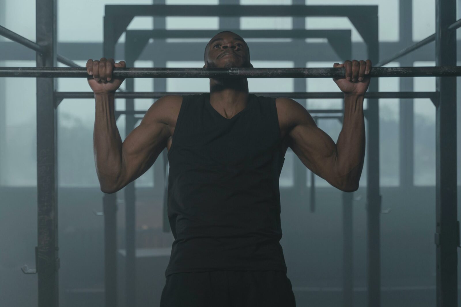 A man in a black sleeveless shirt performs a pull-up on a metal bar in a gym with geometric metal structures in the background, showcasing dedication to men's wellbeing.