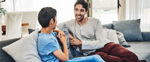 A man and a boy sit on a sofa in a bright living room, smiling and having an R U OK? conversation. The man is wearing a light long-sleeved shirt and red trousers, while the boy wears a blue t-shirt and jeans.