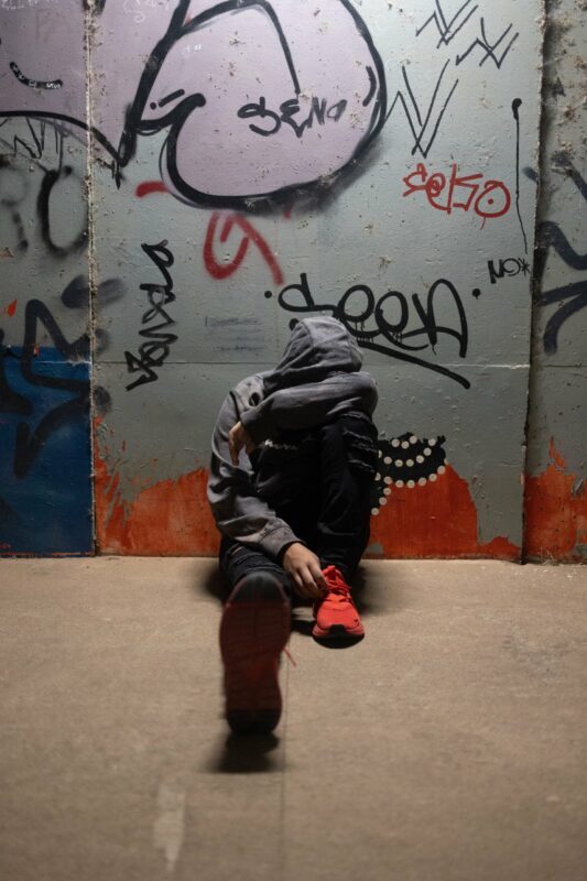 A person in a grey hoodie sits on the floor against a graffiti-covered wall, head lowered and face hidden, with one leg stretched out and wearing bright red shoes. The dimly lit urban scene subtly hints at struggles with self-harm.
