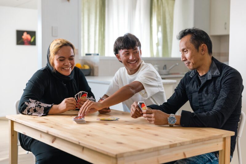 Three people sit around a wooden table in a bright kitchen, smiling and laughing as they play Uno. The lively game reflects the joy and connection found in adolescent relationships, with cards spread out on the table.