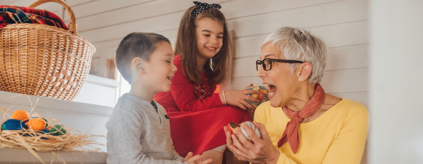 An older woman with short grey hair and glasses excitedly shows a small present to two smiling children on stairs, with a basket and Easter eggs nearby—a joyful moment that highlights what kinship care can look like.