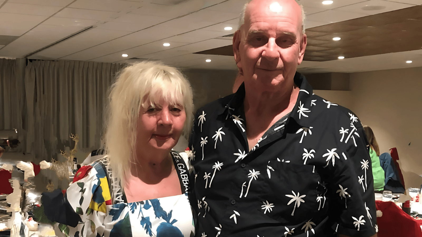 Wayne and Robyn stand closely together at an indoor event. The woman has long blonde hair and a colourful dress, while the man wears a black shirt with white palm trees. They both smile gently at the camera.