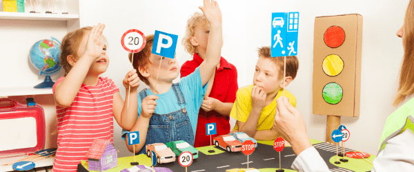 Five children take part in a classroom road safety activity, holding up traffic signs and interacting with a cardboard traffic light and miniature cars on a model road.