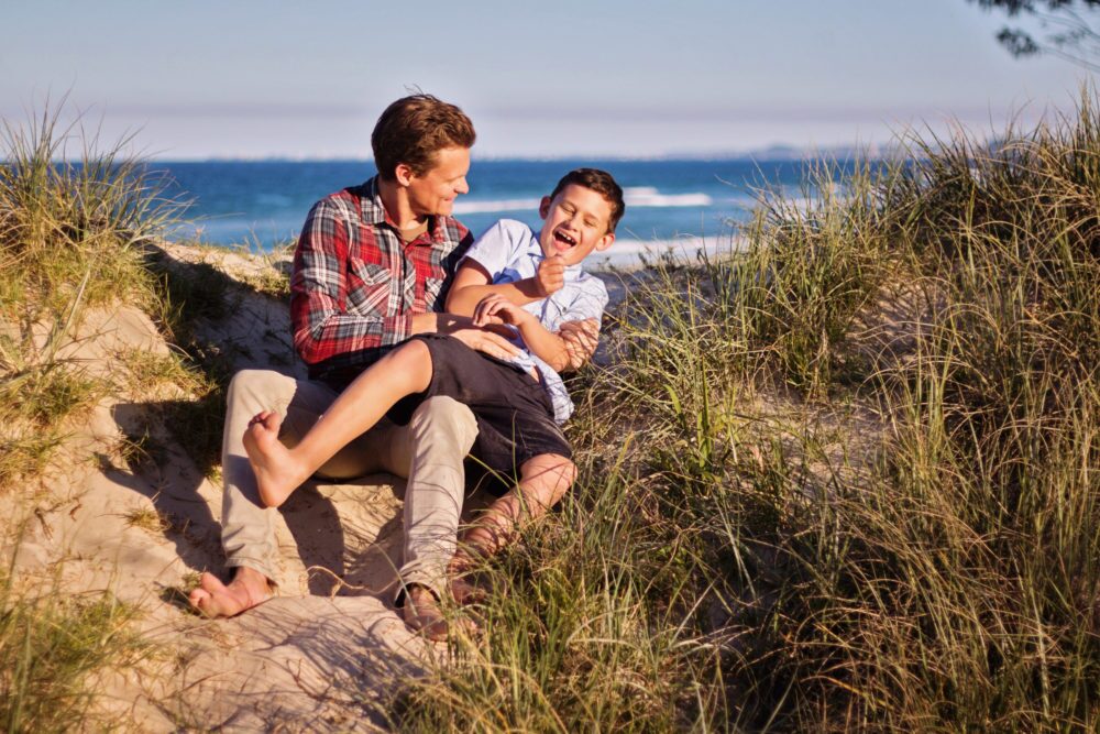 A man and a young boy sit barefoot on sandy dunes, surrounded by grass, laughing and playing together. As the sea glimmers beyond them, one might wonder: how long does it take to become a foster carer and create moments like these?.