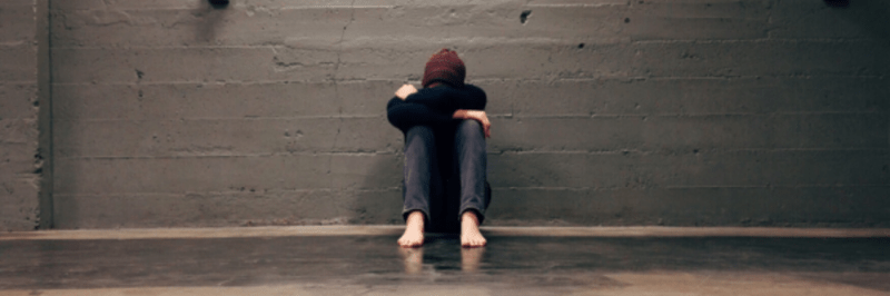 A person sits barefoot on the floor against a grey concrete wall, knees pulled to their chest and arms wrapped around their legs, with their head down, capturing the struggle and isolation often faced by youth homelessness.