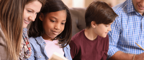 A woman and two children sit together, reading and writing, while an adult next to them holds a pencil. They appear to be enjoying a learning activity at home, celebrating the spirit of Foster and Kinship Carer Week.