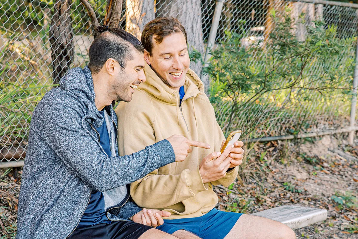 Two men sitting on a bench outdoors, smiling and laughing whilst looking at a mobile. One man is pointing at the phone, and both appear to be enjoying a light-hearted moment together, celebrating LGBTQIA friendship. A chain-link fence is in the background.