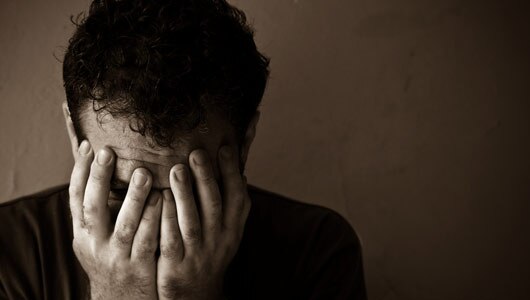 A person with dark curly hair sits against a plain wall, covering their face with both hands in a gesture of stress or sadness. The dim lighting enhances the sombre mood.