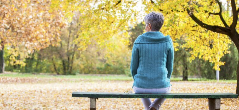 Elderly lady with dementia sitting on a park bench looking at trees.