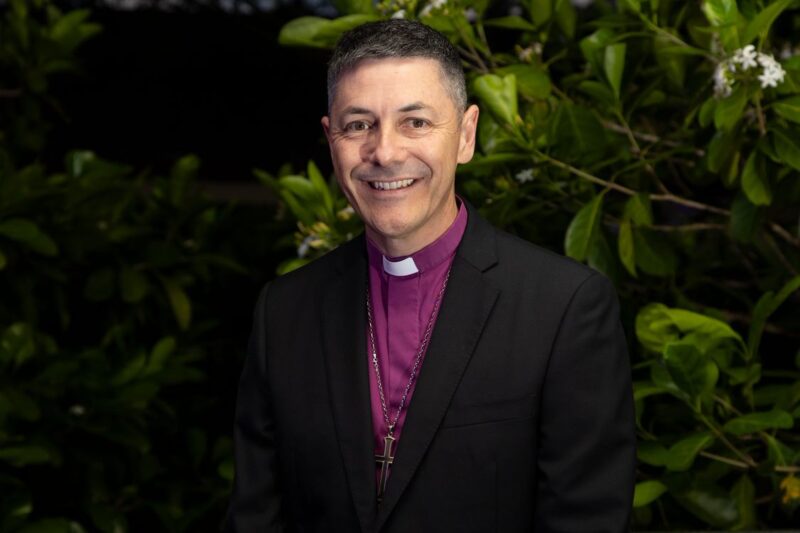 A smiling man wearing a clerical collar, purple clerical shirt, black suit, and cross necklace stands in front of green foliage.