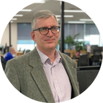 A man with short grey hair and glasses, wearing a grey blazer and light-coloured shirt, stands in a modern office space with desks and computers in the background.