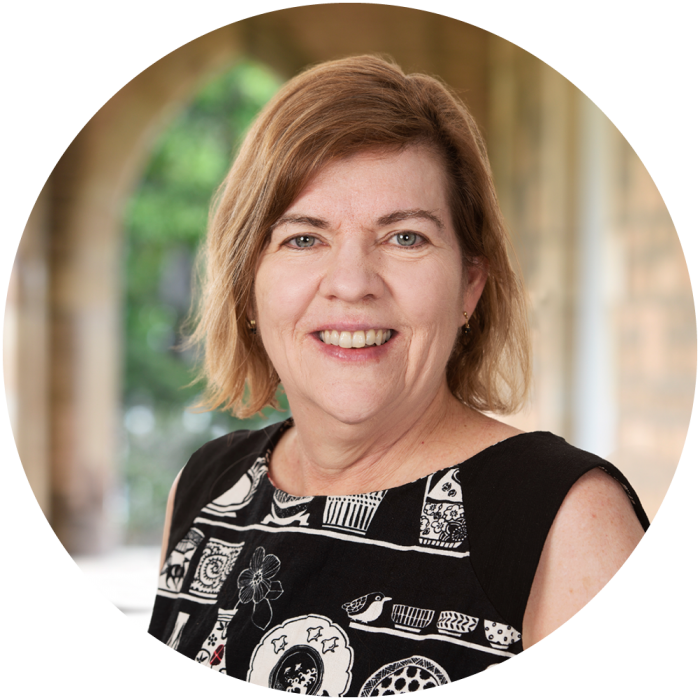 A woman with shoulder-length light brown hair smiles at the camera. She is wearing a black and white patterned top and standing in front of an outdoor background with arches and greenery.