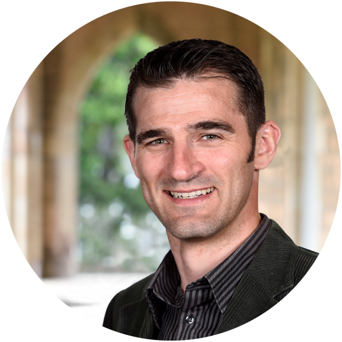 A smiling man with short dark hair, wearing a dark jacket and striped shirt, stands in front of a blurred background featuring an arched stone structure and greenery.