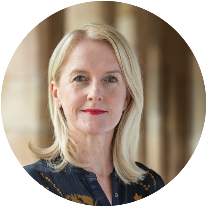 A woman with blonde hair and light skin, wearing a dark blouse with a subtle pattern, stands indoors in soft, natural light, looking confidently at the camera.
