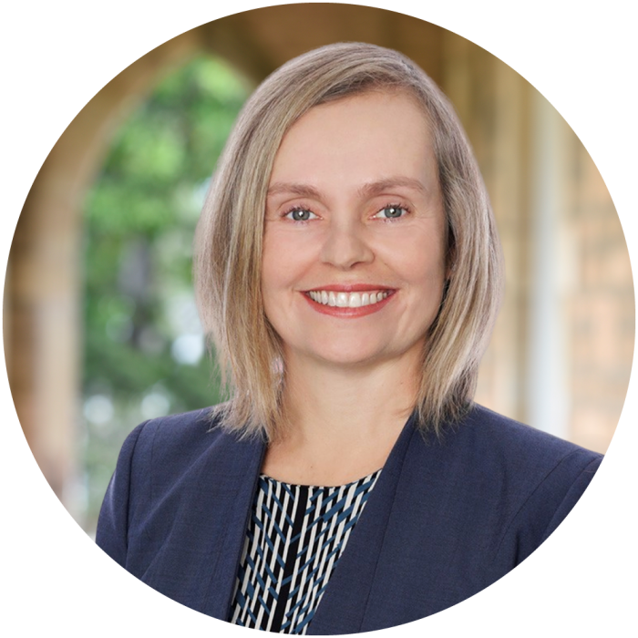 A woman with shoulder-length blonde hair, wearing a navy blazer and patterned top, smiles in front of a blurred background with greenery and an archway.