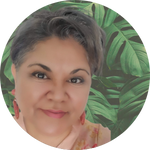 A woman with short dark hair smiles gently at the camera. She is wearing pink earrings and a peach top. The background features large green leaves, creating a tropical feel.