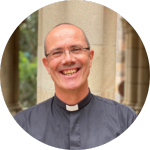 A smiling man wearing glasses and a clerical collar stands outdoors in front of stone columns and greenery.
