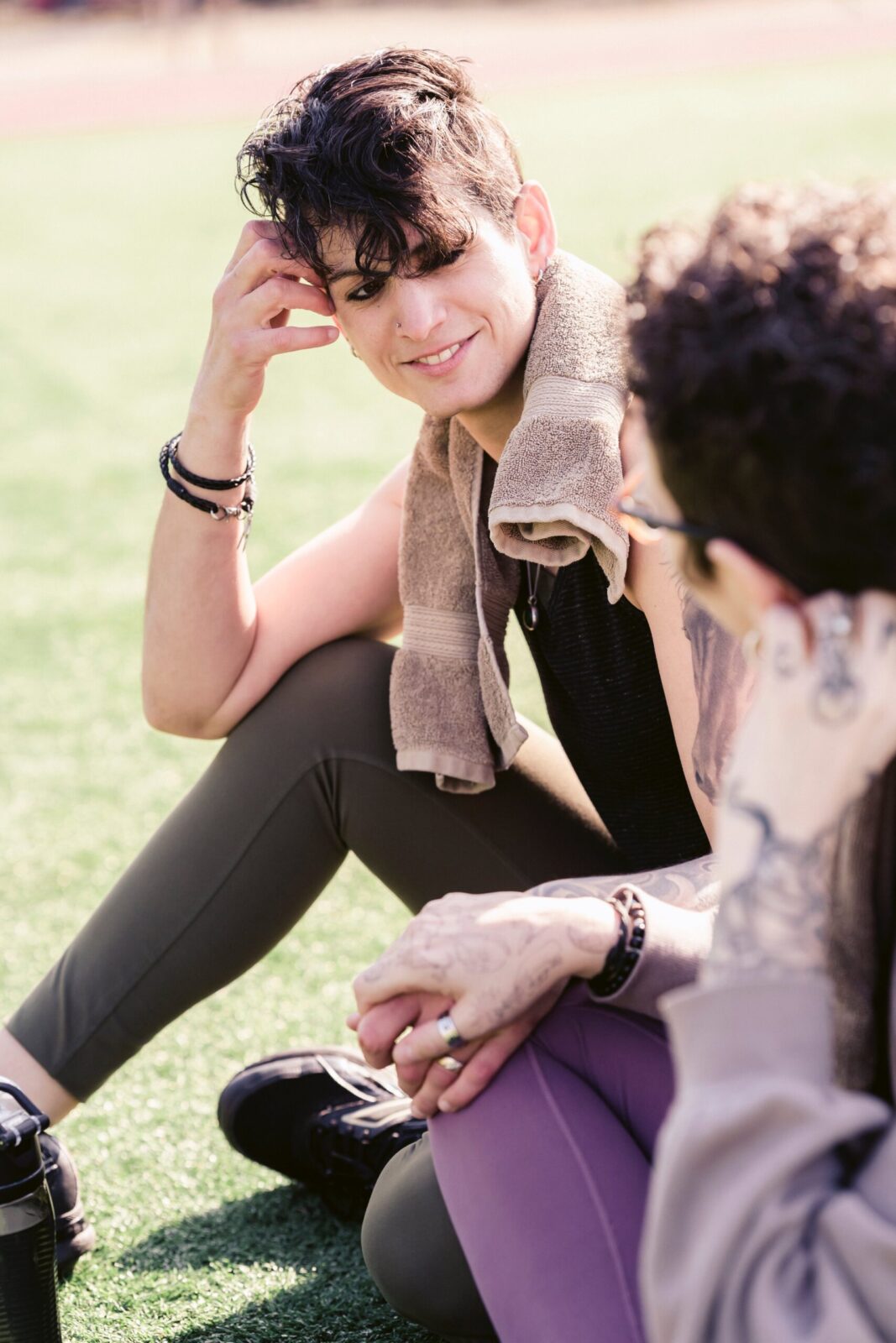 Two people sit on grass, talking and smiling after exercise. One has a towel round their neck and leans forward, while the other listens attentively—showing effective communication as they relax in sports clothing.