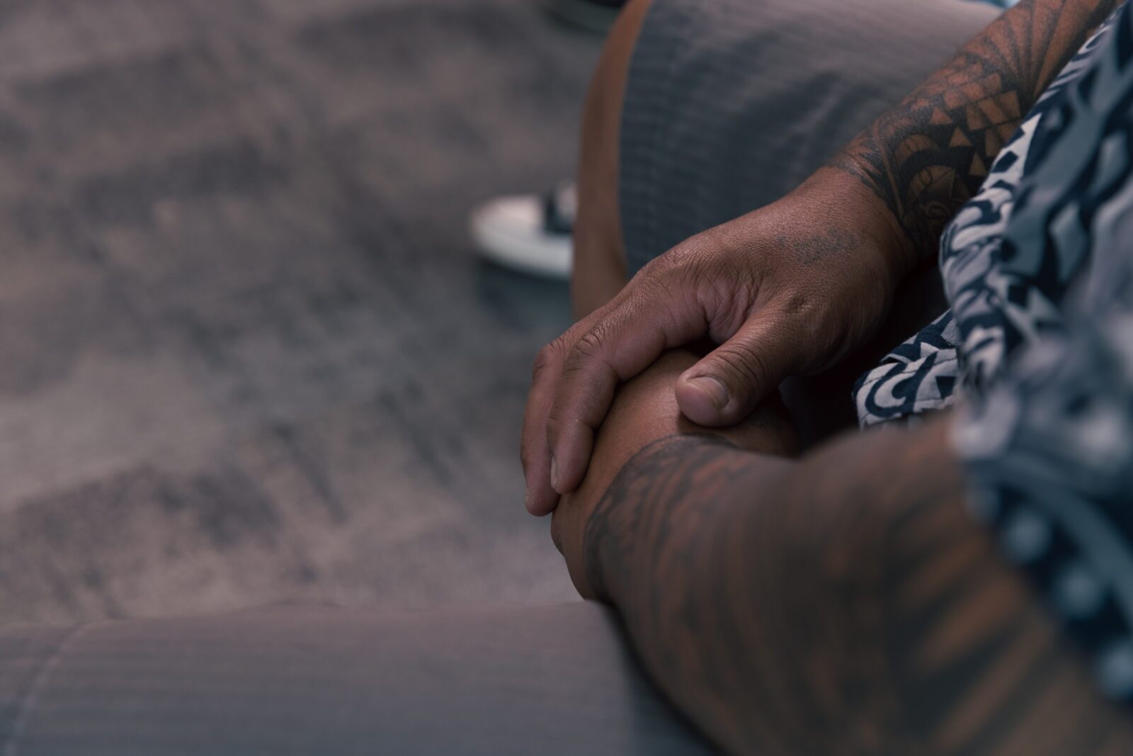 A close-up of a person’s hands resting on their lap. The person, whose tattooed arms hint at stories within the aggression cycle, is wearing shorts and a patterned shirt. The blurred background reveals part of a trainer and a textured floor.