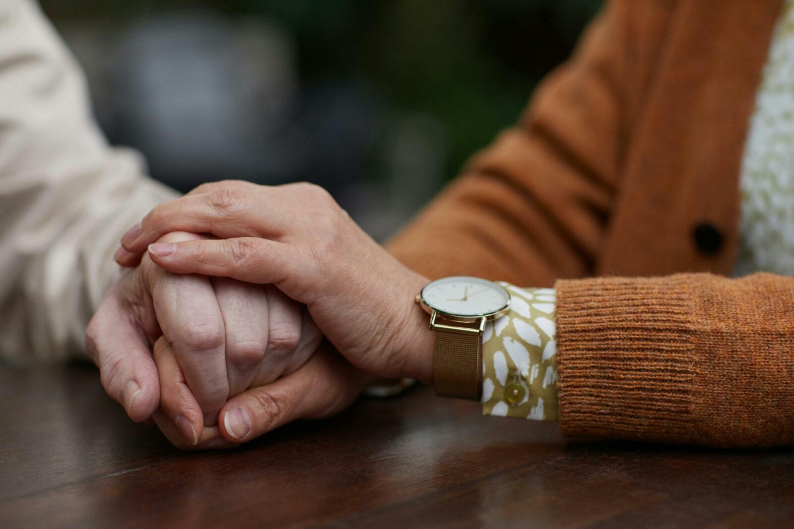 Two people hold hands on a wooden table, one wearing a brown jumper and a gold watch, showing relationship skills as they offer comfort and support to the other.