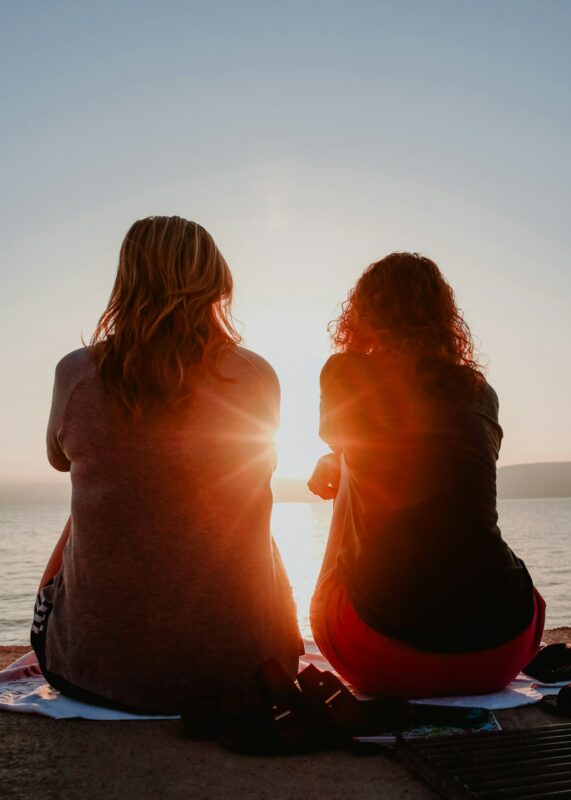 Two people sit side by side on a jetty, facing a calm body of water during sunset. As sun rays shine between them, they share a quiet moment together, finding peace and comfort away from stressful situations.