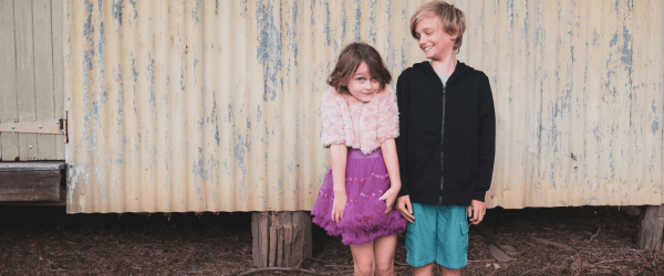 Two children, both in care and keeping siblings together, stand before a yellow corrugated wall. The girl smiles shyly in a pink top and purple skirt, whilst her brother in a black hoodie and teal shorts looks at her, smiling.