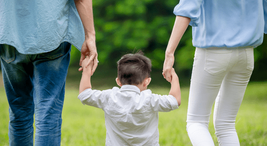 A young child holding hands with two adults, walking together in a grassy outdoor area with green trees in the background—a heart-warming moment of reunification, photographed from behind with the child in the centre.