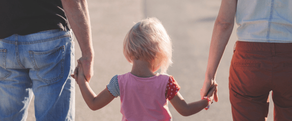 A young child with blonde hair holds hands with two adults, one on each side, as they walk outdoors—capturing a tender moment that highlights the many reasons to foster children and support their development.