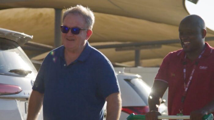 Two men smiling outdoors near parked cars; one wears a blue shirt and sunglasses, the other wears a red shirt and lanyard, with sunlight casting shadows on the scene.