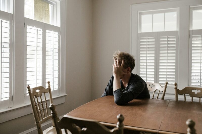 A person sits alone at a wooden table in a bright room with white shutters, resting their head on one hand, lost in thinking patterns that suggest stress or deep contemplation.