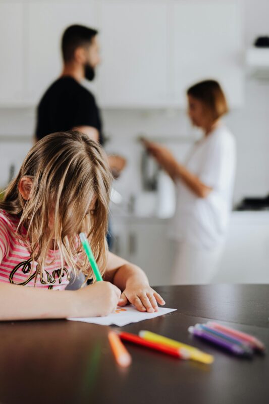 Child colouring in at a table whilst parents experience parental burnout.
