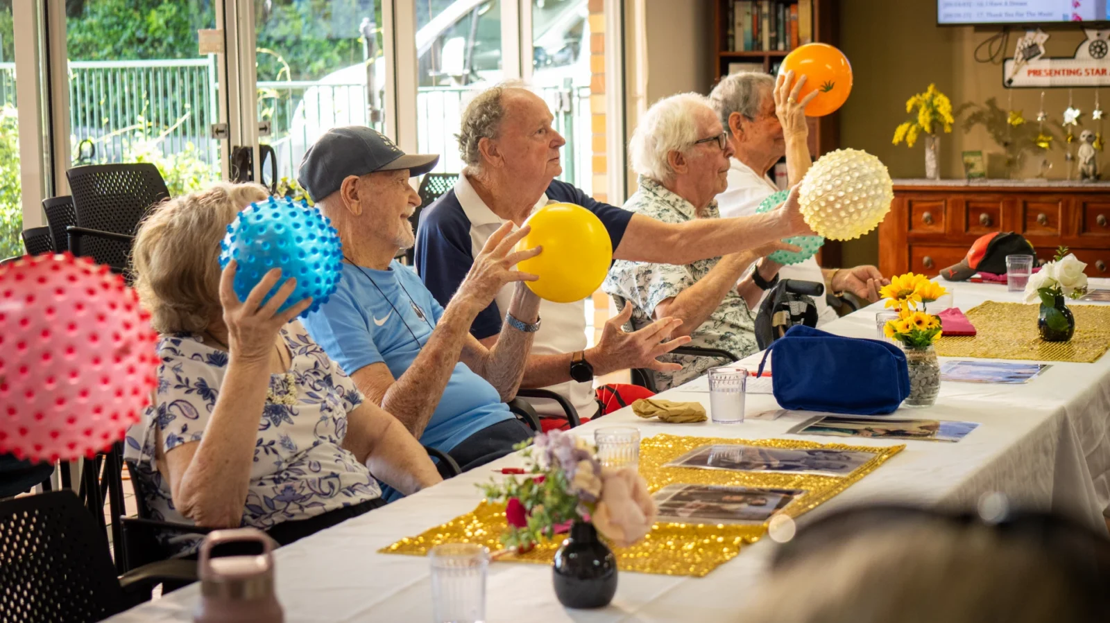 A group of elderly people sit at a table in a bright Anglicare Southern Queensland room, each holding a colourful ball and participating in a group activity. The table is decorated with flowers and golden runners.