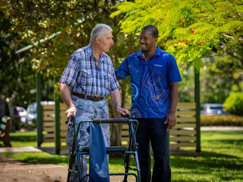 Anglicare Southern Queensland aged care worker supporting elderly client to walk with her wheelie walker.