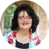 A woman with dark hair and red glasses smiles, wearing a floral jacket over a black top and a pearl necklace. The background is softly blurred, showing an outdoor corridor with greenery.