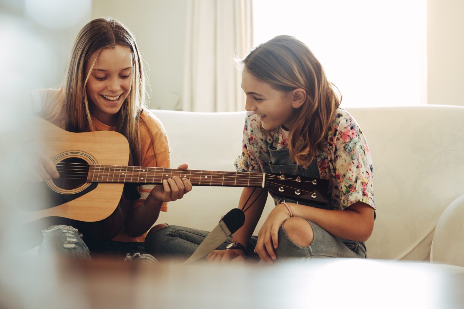 Two teenage girls sitting on a sofa, smiling and enjoying playing an acoustic guitar together in a sunlit room. Their happiness hints at the warm bonds that can form in supportive homes—what kind of foster care makes moments like this possible?.